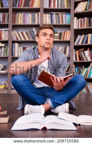 Waiting for inspiration. Thoughtful young man holding note pad and touching his chin with pen while sitting against bookshelf