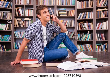 Talking with friend. Happy young man holding hands on chin and smiling while sitting against bookshelf
