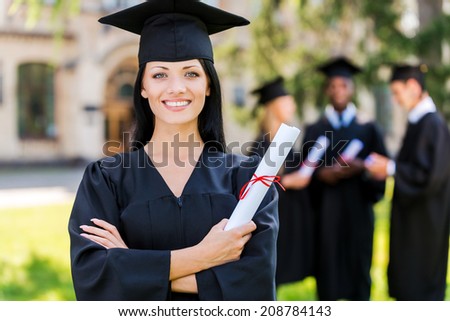 Happy to be graduated. Happy young woman in graduation gowns holding diploma and smiling while her friends standing in the background