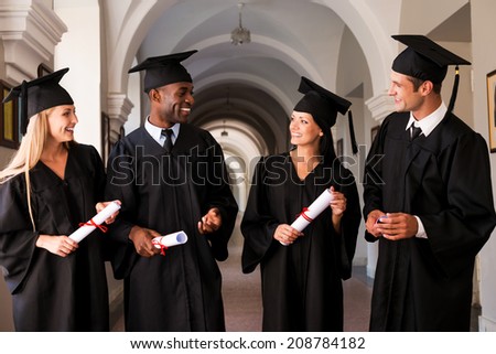 Talking about bright future. Four college graduates in graduation gowns walking along university corridor and talking