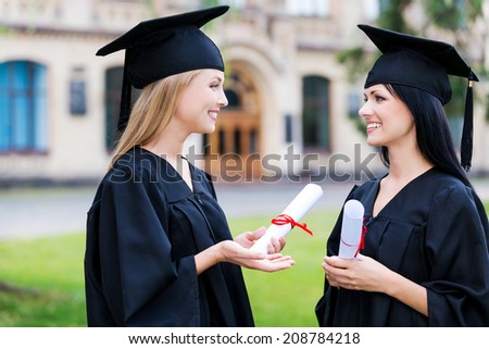 Discussing ceremony. Two happy young women in graduation gowns holding diplomas and talking