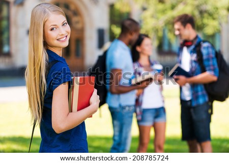 My future is in my hands. Beautiful young woman holding books and smiling while standing near university building and with her friends chatting in the background