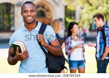 Enjoying university life. Handsome young African man holding books and smiling while standing against university with his friends chatting in the background