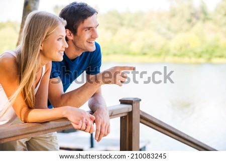 Look over there! Beautiful young loving couple leaning at the wooden railing and smiling while man pointing away