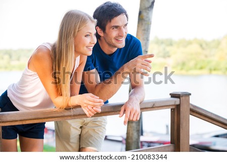 What is that? Beautiful young loving couple leaning at the wooden railing and smiling while man pointing away