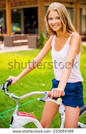 Ready to ride my new bike! Beautiful young blond hair woman smiling and looking at you while sitting on her bicycle outdoors