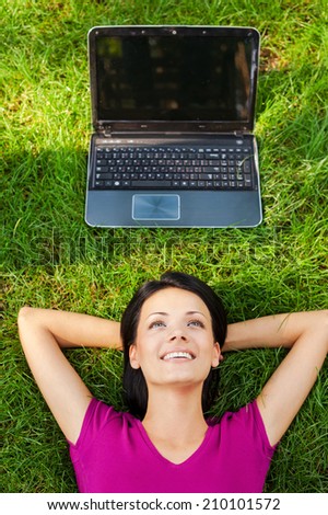 Living in digital age. Top view of beautiful young woman lying in grass and smiling while while laptop laying upon her head