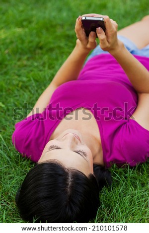 Staying in touch with friends. Attractive young woman typing message on mobile phone and smiling while lying in grass