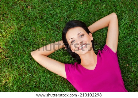 Relaxing in grass. Top view of beautiful young woman holding hands behind head and smiling to you while lying on the green grass
