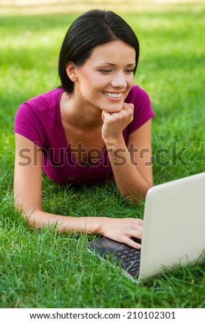 Surfing the net outdoors. Attractive young woman working at laptop and smiling while lying in grass
