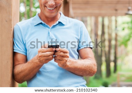 Examining his new smart phone. Cheerful mature man holding mobile phone and smiling while standing outdoors