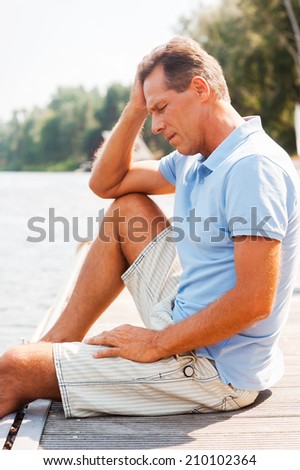 Depressed man. Side view of depressed mature man touching head with hand and keeping eyes closed while sitting at the quayside