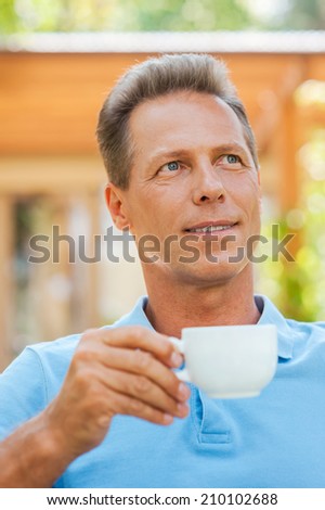 Day dreaming with cup of coffee. Cheerful mature man drinking coffee and smiling while sitting outdoors with house in the background