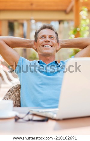 Dreaming of vacation. Relaxed mature man holding hands behind head and smiling while sitting at the table outdoors with laptop on it