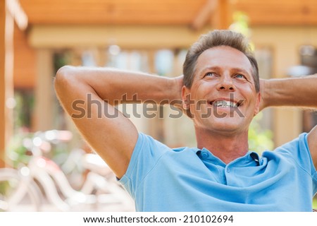 Happy day dreamer. Relaxed mature man holding hands behind head and smiling while outdoors with house in the background