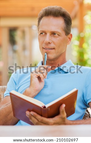 Waiting for inspiration. Thoughtful mature man holding note pad and touching his chin with pen while sitting outdoors