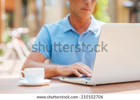 Surfing the net in cafe. Close-up of confident mature man writing something in his note pad and smiling while sitting at the table outdoors with house in the background
