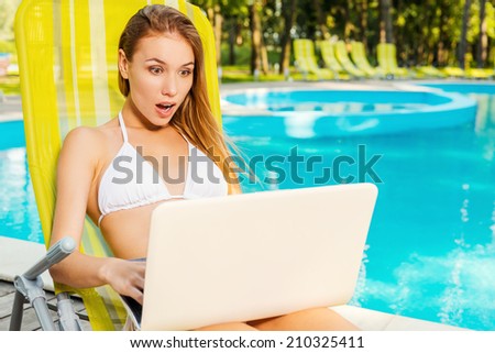 It is just unbelievable! Beautiful young woman in white bikini looking at laptop and keeping mouth open while sitting at the deck chair by the pool