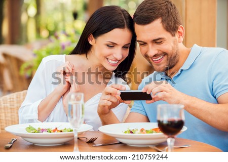 I want to share this picture with friends. Happy young loving couple taking pictures of their food and smiling while relaxing in outdoors restaurant together