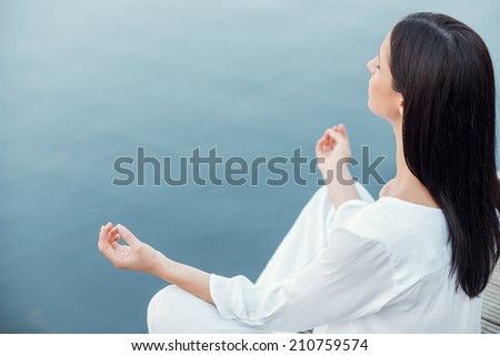 In peace with universe. Top view of beautiful young woman in white clothing meditating while sitting on the wooden quayside