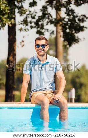 Spending his leisure time poolside. Handsome young man in sunglasses sitting by the pool and smiling