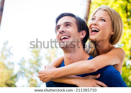 Happy to be together. Low angle view of happy young loving couple standing outdoors together while woman hugging her boyfriend from back and smiling