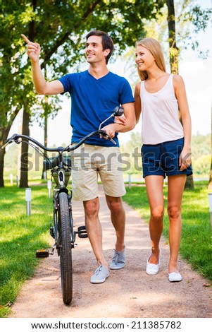 Couple with bicycle. Beautiful young smiling couple walking with bicycle in park while man pointing away and smiling