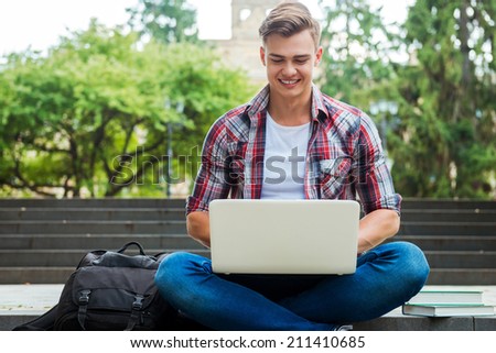 Surfing the net outdoors. Happy male student working on laptop and smiling while sitting at the outdoors staircase with books and backpack laying near him