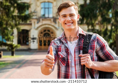 Thumb up for success! Handsome male student showing his thumb up and smiling while standing outdoors with university building in the background