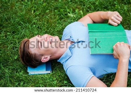 Day dreaming with his favorite book. Top view of happy young man holding a book and smiling while lying on grass with laptop