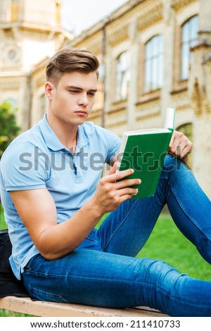 Confident bookworm. Confident male student reading book while sitting on the bench and in front of university building