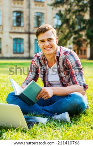I love studying. Cheerful male student reading book and smiling while sitting on the grass and in front of university building