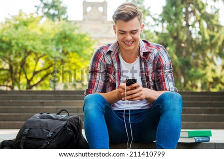 Enjoying his favorite music. Happy male student listening to MP3 Player and smiling while sitting at the outdoors staircase with books and backpack laying near him
