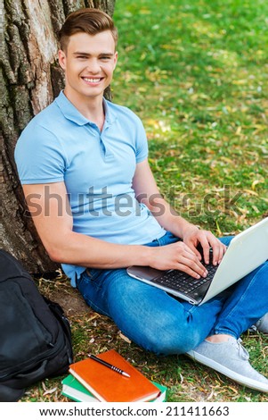 Finding a peaceful place for studying. Top view of confident male student working on laptop and smiling while sitting on the grass and leaning at the tree