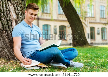 Dedicated to studying. Confident male student reading book and writing something in note pad while sitting on the grass and in front of university building