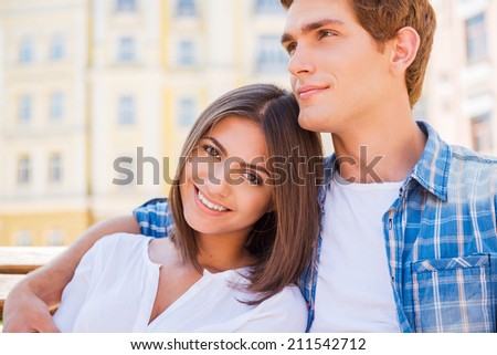 Enjoying their time together. Beautiful young loving couple sitting on the bench together while woman looking at camera and smiling