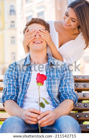 Guess who? Beautiful young woman covering eyes of her boyfriend and smiling while man sitting on the bench and holding red rose