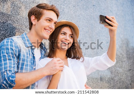 Catching the bright life moments. Beautiful young loving couple making selfie with smart phone and smiling while leaning at the wall