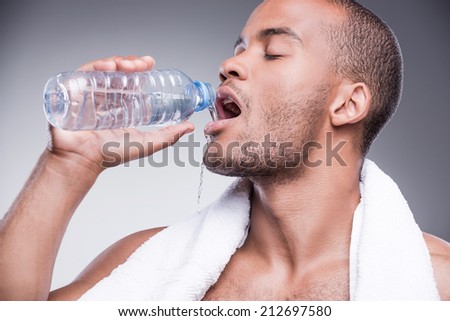 Refreshing after training. Young shirtless African man holding bottle with water and smiling while standing against grey background
