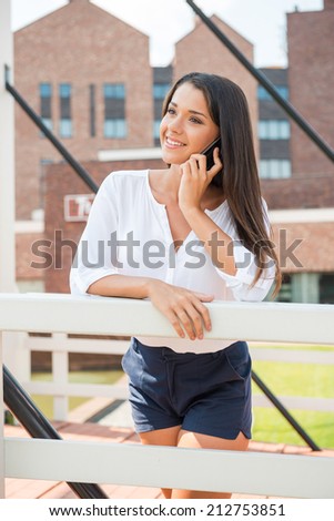 Talking with friend. Beautiful young woman talking on the mobile phone and smiling while standing outdoors