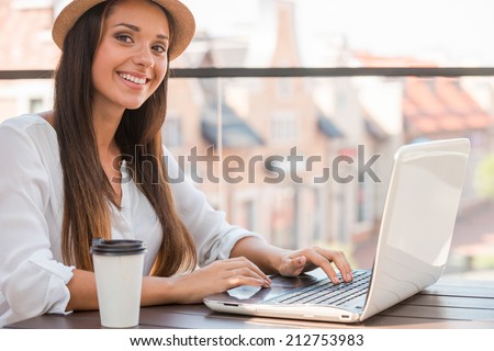 Surfing the net outdoors. Beautiful young woman in funky hat working on laptop and smiling while sitting outdoors