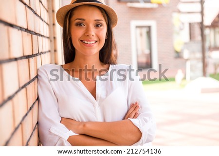 Beautiful and trendy. Beautiful young woman in hat keeping arms crossed and smiling while leaning at the wall outdoors