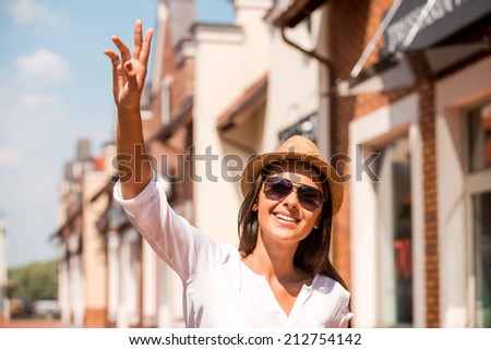 Meeting friend. Beautiful young woman in hat and sunglasses waving to someone and smiling while standing outdoors