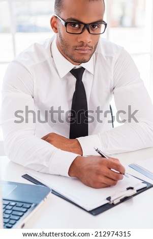 Confident businessman at work. Top view of confident young African man in shirt and tie writing something at clipboard and looking at camera while sitting at his working place