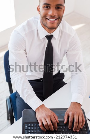 Businessman at work. Top view of confident young African man in shirt and working on laptop and smiling while sitting at his working place