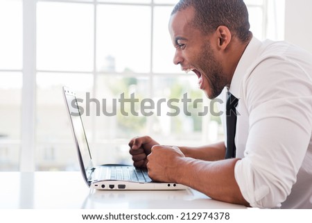 Stressed office worker. Side view of furious young African man in shirt and tie shouting while looking at laptop while sitting at his working place