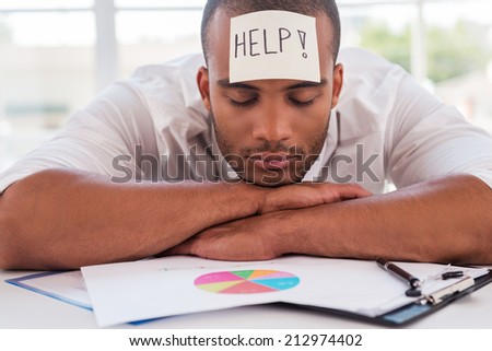 I need help. Frustrated young African man in formalwear with adhesive note on his forehead leaning his head at the table