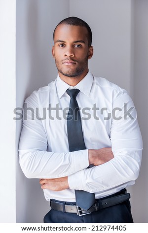 Successful businessman. Handsome young African man in shirt and tie looking at camera and keeping arms crossed while leaning at the wall