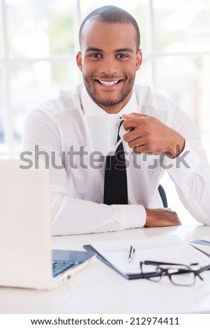 Coffee break. Handsome young man in shirt and tie drinking coffee and smiling while sitting at his working place