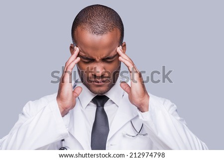 Feeling awful headache. Depressed African doctor touching his head with hands and keeping eyes closed while standing against grey background